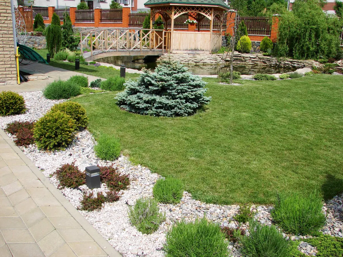 Landscaped yard with a lawn, shrubs in a white stone border, a paver walk, and a wooden gazebo by a pond