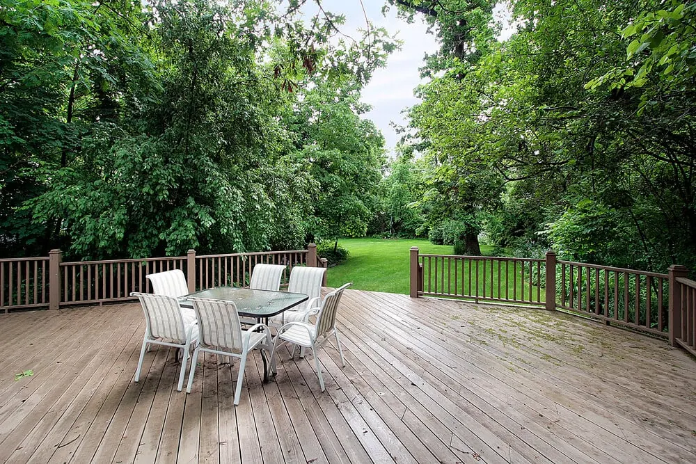 Wooden deck with a glass table and white chairs looking out over a lawn bordered by tall trees