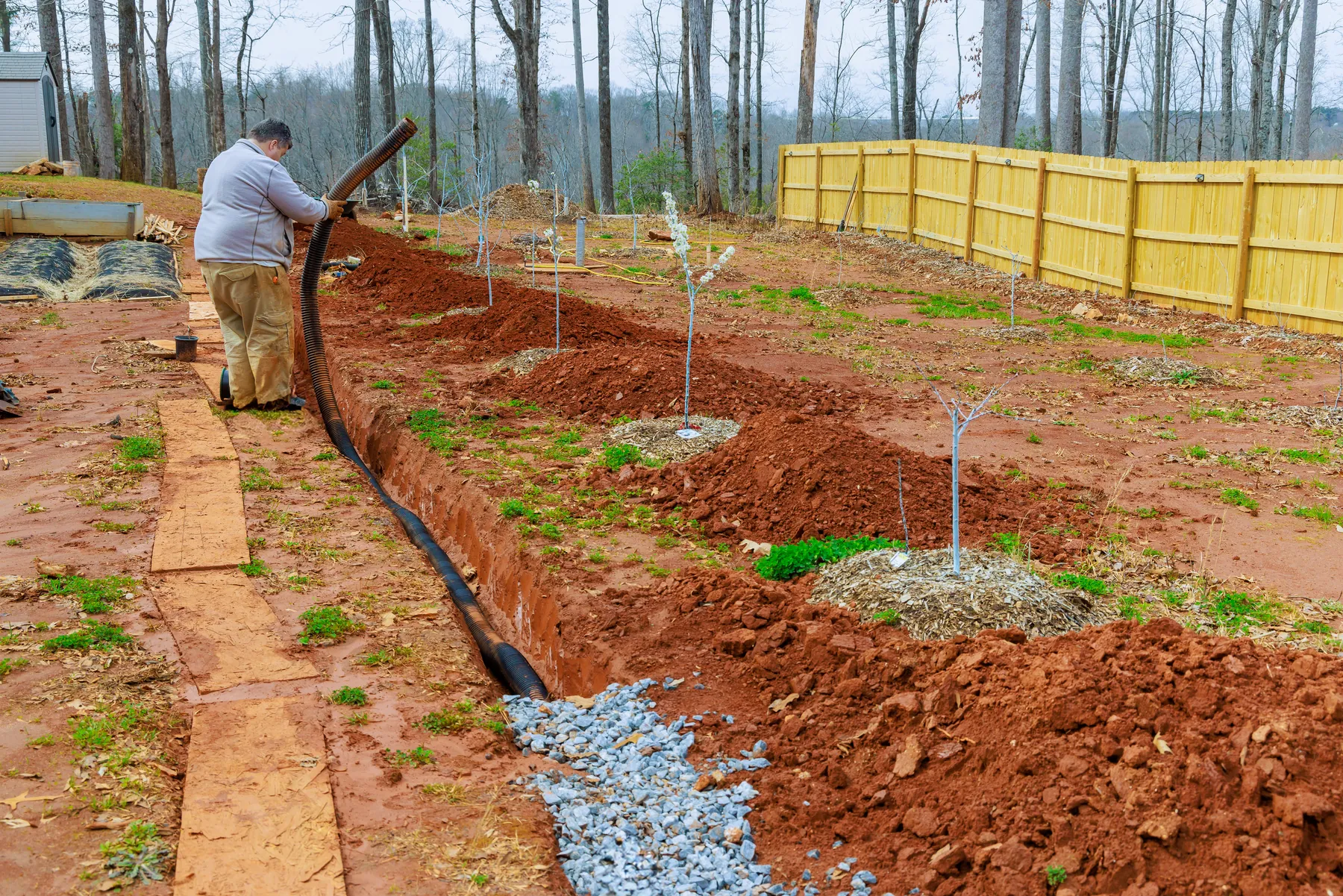 Black perforated drain pipe being laid in a gravel-lined trench across a yard