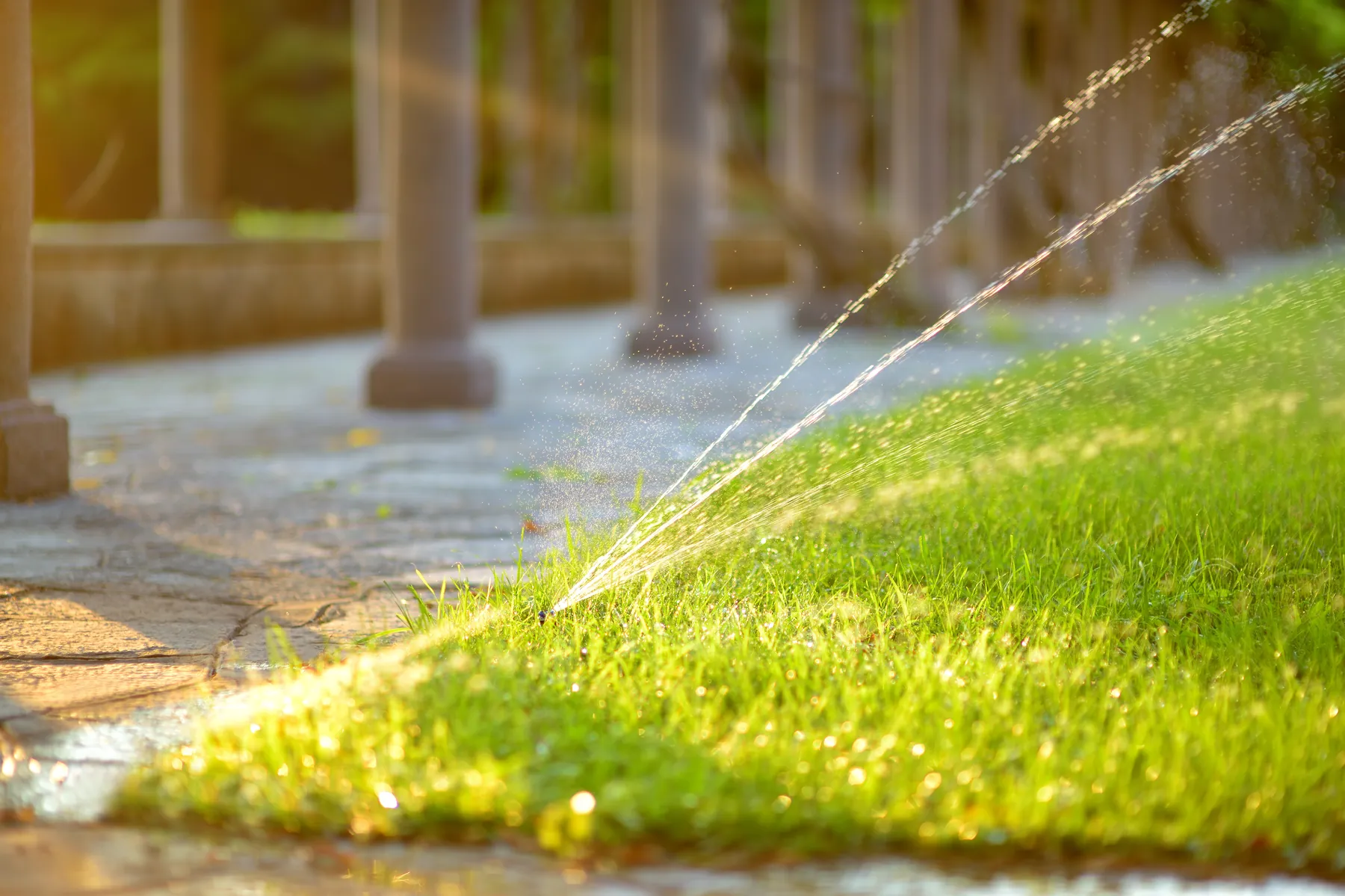Sprinkler spraying a home lawn beside a paved patio and columns at sunset
