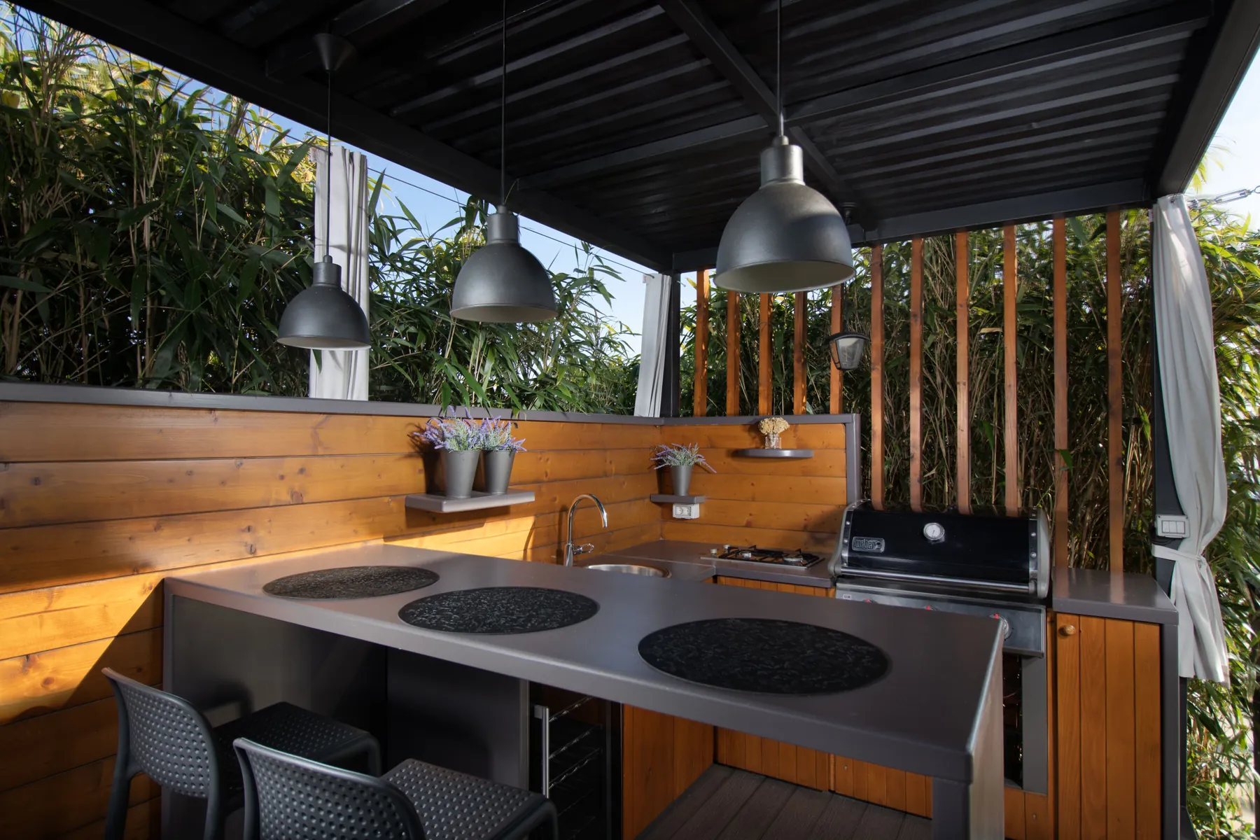 Covered outdoor kitchen with a stainless grill, gray counters and pendant lights under a solid roof