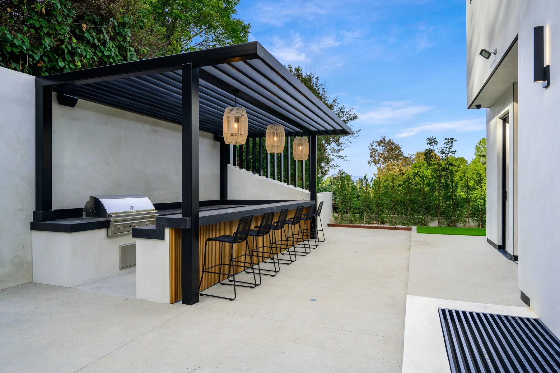 Large outdoor kitchen with a built-in grill, bar counter and stools under a slatted roof