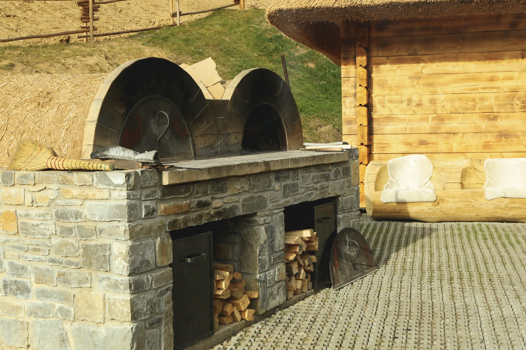 Built-in stone oven with arched openings and stacked firewood beside a wooden house