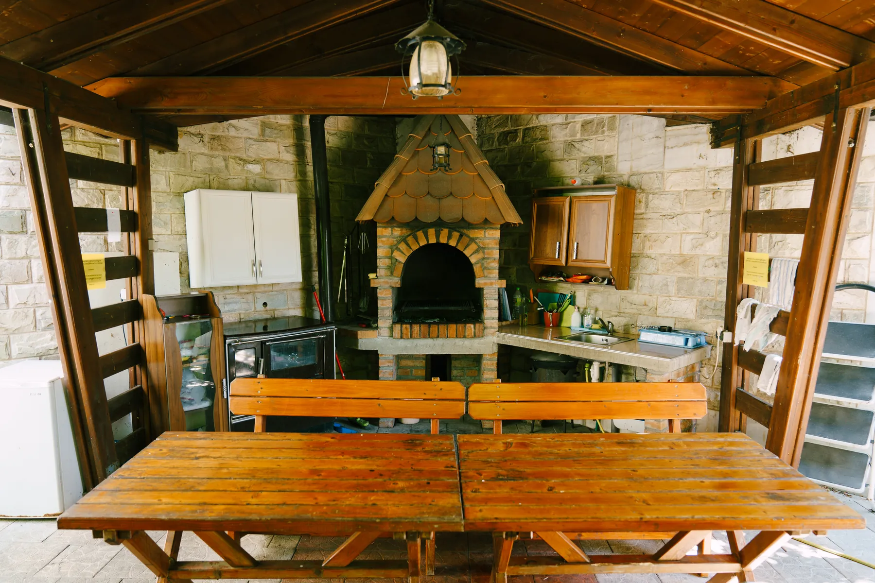 Wooden outdoor kitchen pavilion with a stone barbecue, cabinets and a long plank table