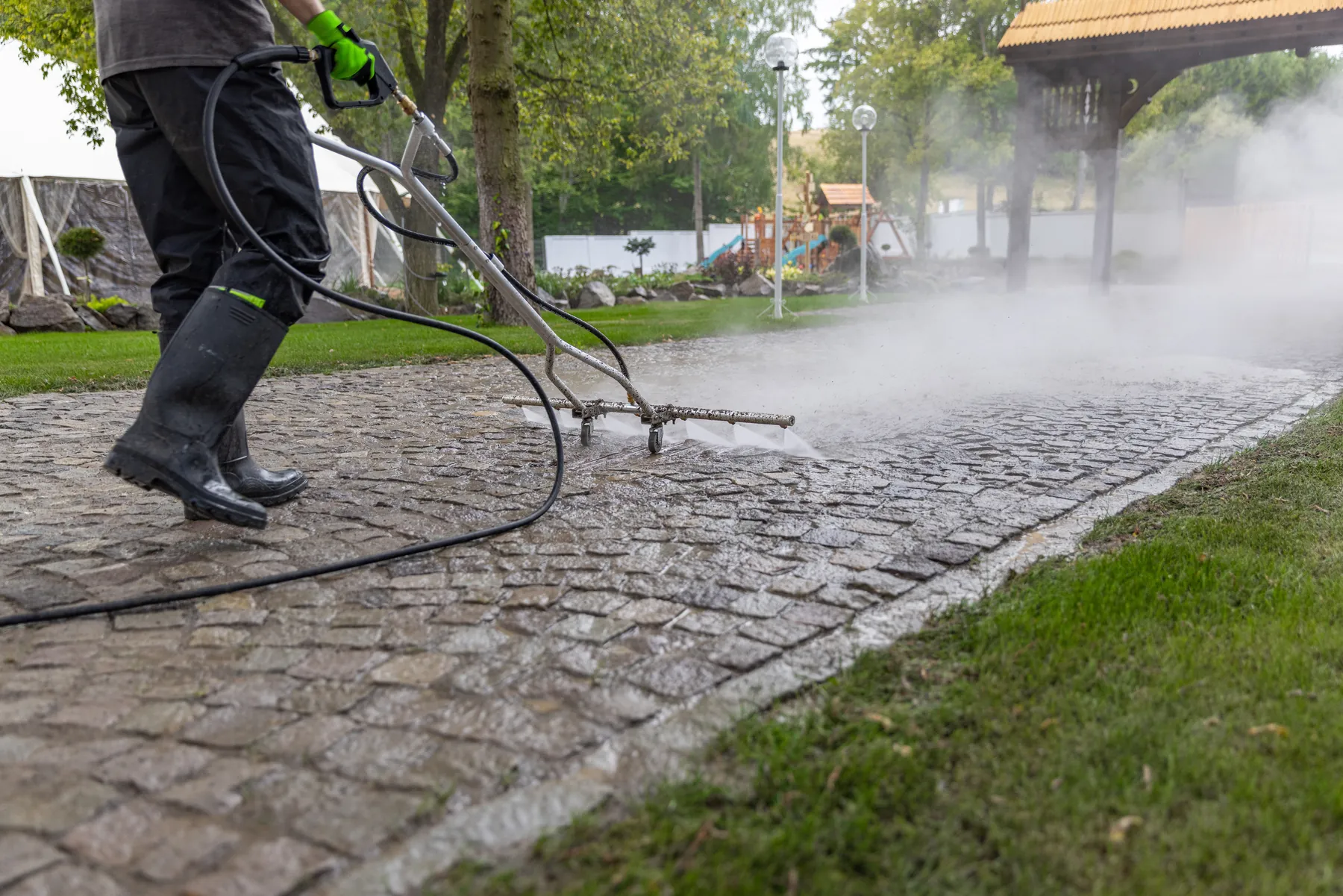 Pressure washer wand cleaning a cobblestone driveway beside a lawn